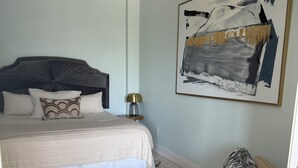 Een slaapkamer met een bed, nachtkastje, lamp, stoel en een schilderij aan de muur.