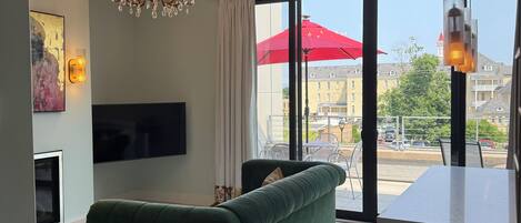 A modern living room with a green sofa, a chandelier, a flat-screen TV, and a large window with a view of buildings and a red umbrella.