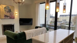 A modern living room with a green sofa, a flat-screen TV, and a large window offering a city view. - Asylum 58 Condo @ Grand Traverse Commons (Traverse City)