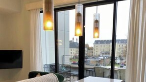 A modern hotel room with a large window, a flat-screen TV, a sofa, a dining table, and pendant lights. - Asylum 58 Condo @ Grand Traverse Commons (Traverse City)
