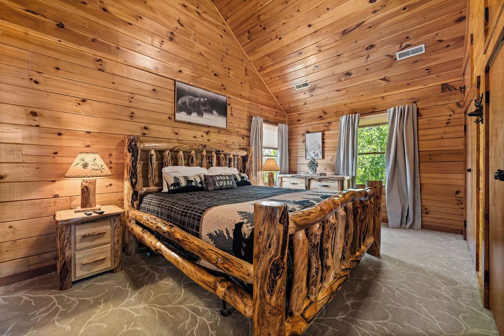 A wooden cabin bedroom with a rustic bed frame, a nightstand, a lamp, and a window with curtains.