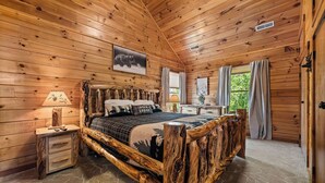A wooden cabin bedroom with a rustic bed frame, a nightstand, a lamp, and a window with curtains. - Mountain Paradise (Sevierville)