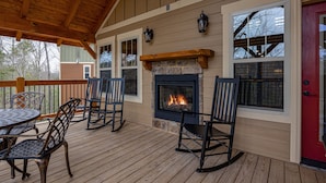 A covered deck with a stone fireplace, wooden railing, and metal chairs. - Serenity Peak (Gatlinburg)