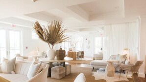 A spacious, well-lit living area with a modern design, featuring a central seating arrangement, a large decorative plant, and a dining area in the background.