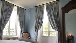 Ein Schlafzimmer mit einem großen Bett, zwei mit Vorhängen versehenen Fenstern, einem Sessel, Holzboden und einem Spiegel.