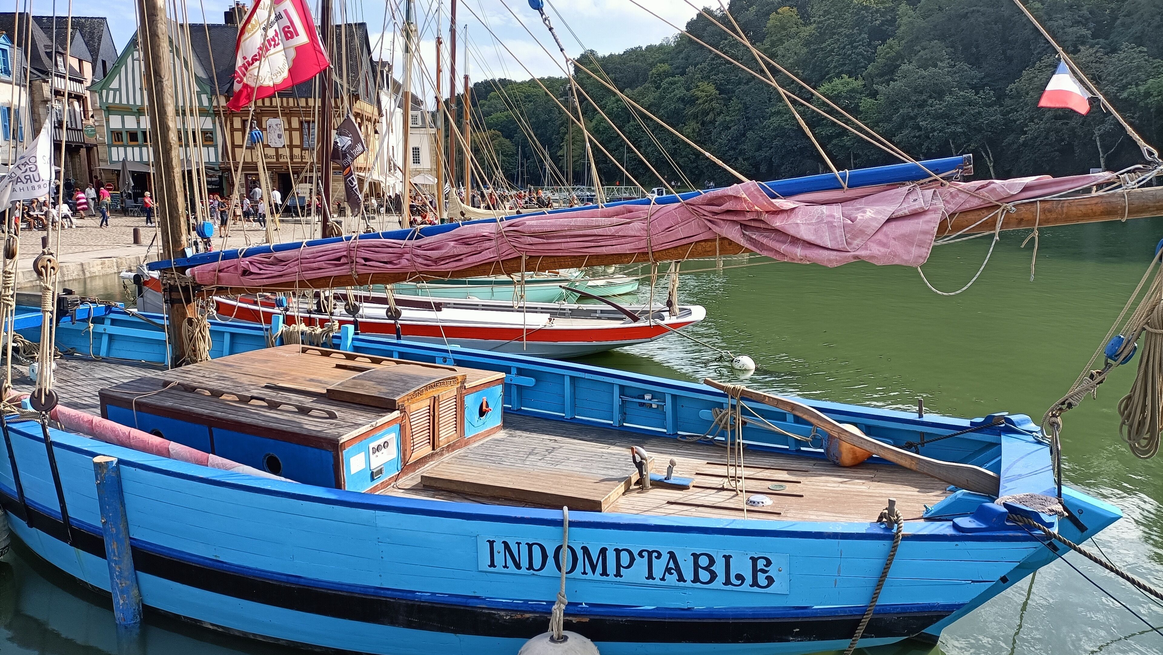 A blue boat named 'INDOMPTABLE' docked at a harbor with a crowd and buildings in the background.