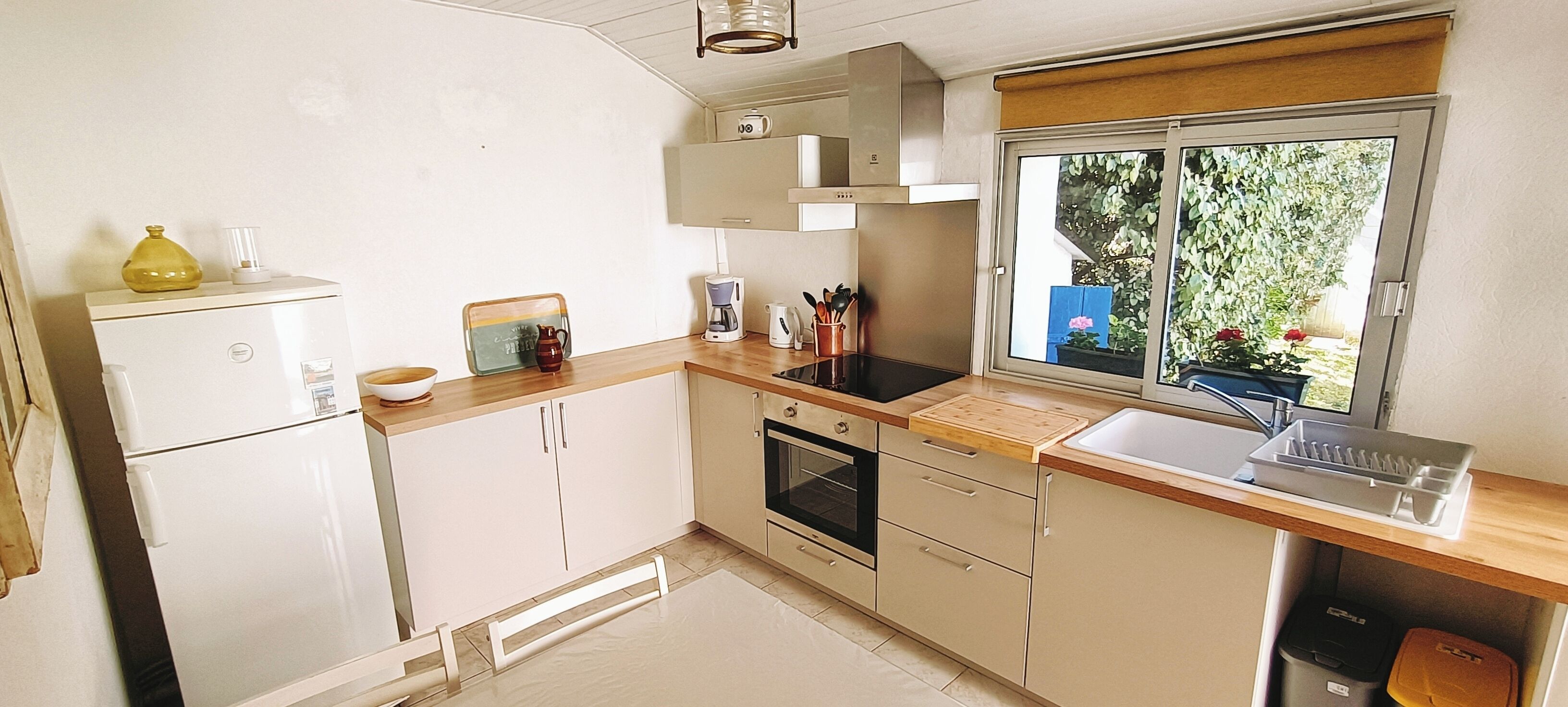 Een kleine keuken met een koelkast, oven, gootsteen en raam met uitzicht op een tuin.