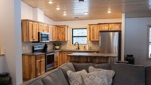 A modern kitchen with wooden cabinets, stainless steel appliances, and a grey sofa with patterned cushions. - NEW 5BR Cabin (Wetmore)