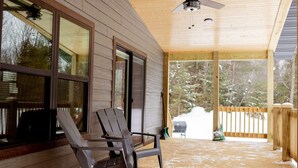 A covered porch with two Adirondack chairs, a ceiling fan, and a view of a snowy landscape. - NEW 5BR Cabin (Wetmore)