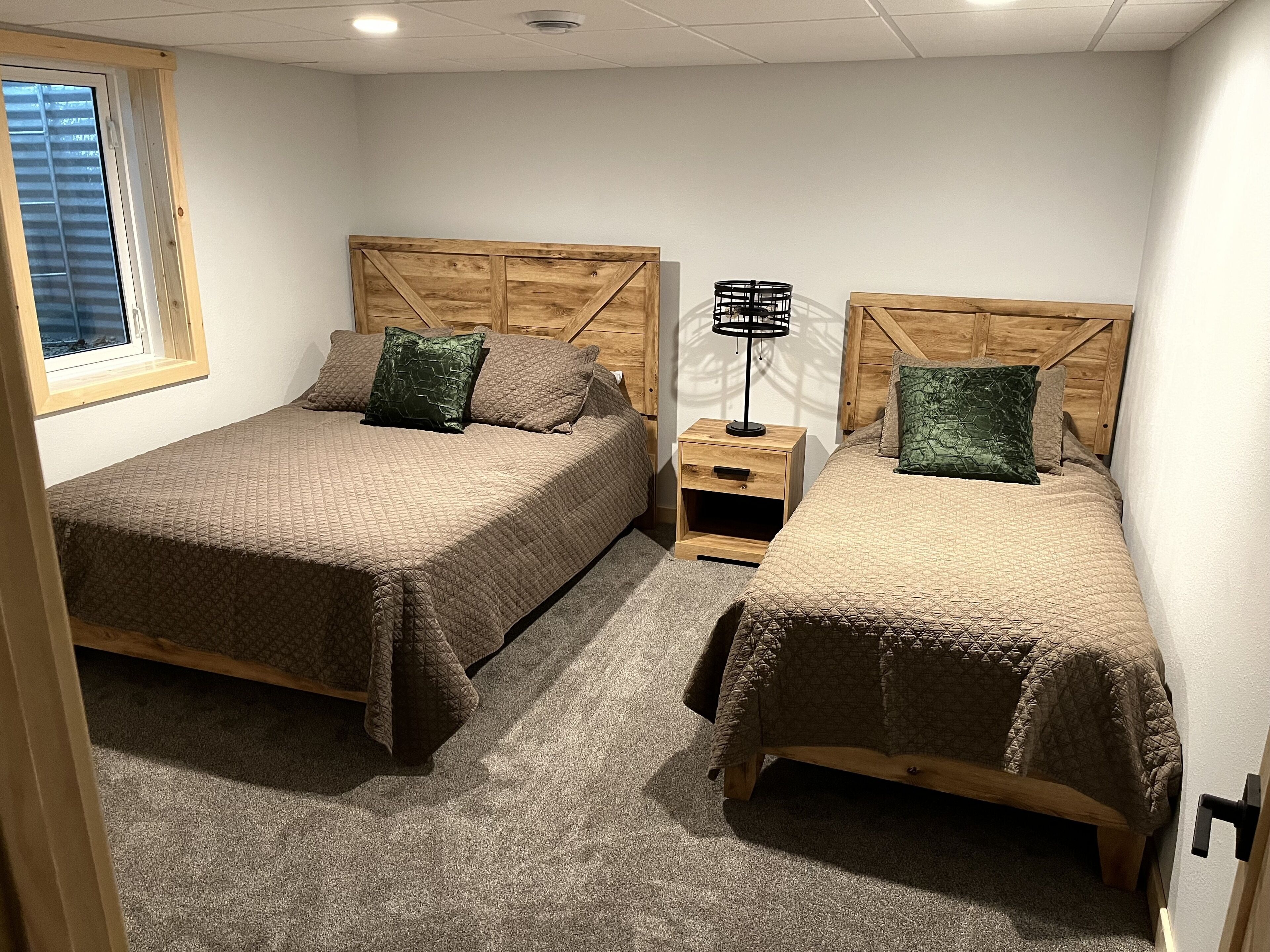 Two twin beds with wooden headboards, a small bedside table with a lamp, and a window with blinds.