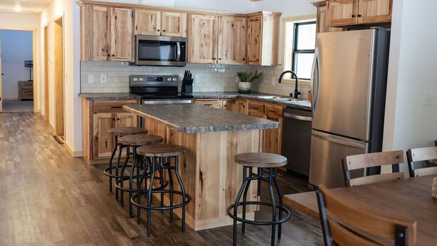 A modern kitchen with wooden cabinets, a stainless steel refrigerator, and a central island with stools. - NEW 5BR Cabin (Wetmore)