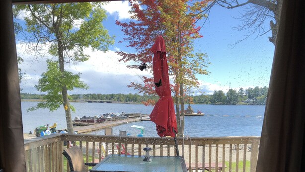 A view from a window overlooking a lake with a dock, a table with chairs, and a red umbrella. - Grace Land Cottages (West Nipissing)