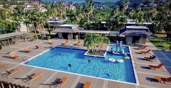 A swimming pool with lounge chairs and umbrellas, surrounded by palm trees and buildings.
