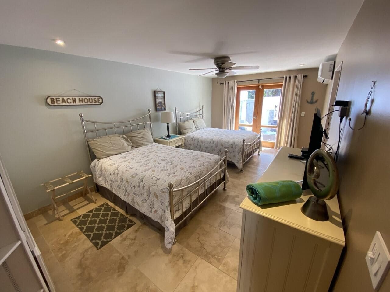 A bedroom with two beds, a ceiling fan, a small desk, and a sign reading "BEACH HOUSE".