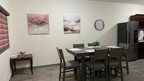 Een eetruimte met een tafel voor vier personen, omringd door stoelen. Er hangen twee schilderijen aan de muur, een klok en een bijzettafeltje met een bloevas. In de achtergrond is een koelkast zichtbaar.
