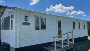 Een witte mobiele woning met een bord waarop 'Summer Breeze Deluxe' staat. Het heeft een veranda met een houten leuning en een trap die naar de ingang leidt.