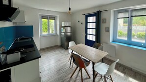 A kitchen with a dining table and chairs, a refrigerator, and a window with blinds. - Near Paimpol, Apartment 200m from Bréhec Beach (Plouézec)