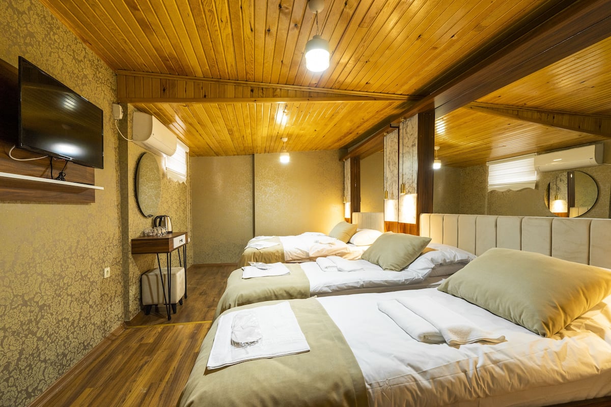 A hotel room with wooden ceilings, two beds with pillows and blankets, a flat-screen TV mounted on the wall, and a small table with a lamp.