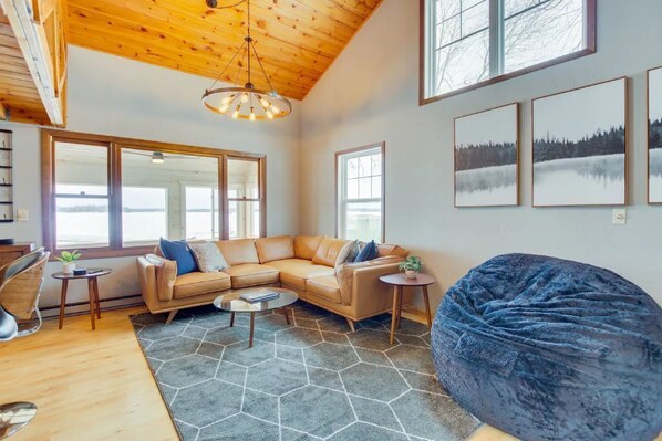 A spacious living room with a large sectional sofa, a round coffee table, and a bean bag chair. There are large windows providing natural light and a view of the outdoors.