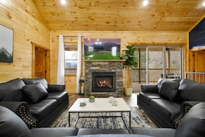 A cozy living room with a stone fireplace, black leather sofas, and a television mounted above it.