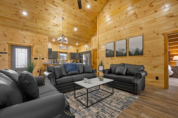 A cozy log cabin interior with a living room featuring two black leather sofas, a coffee table, and a kitchen area with modern appliances.