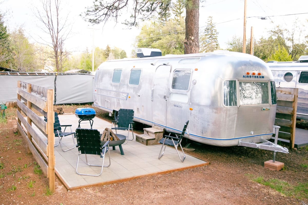 Ein silberner Airstream-Wohnwagen mit einer überdachten Terrasse, grünen Klappstühlen und einem Grill.