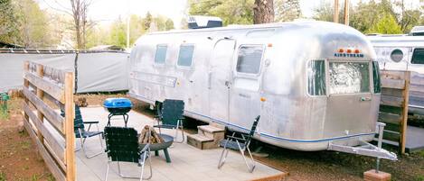 Un rimorchio Airstream argentato con un patio coperto, sedie pieghevoli verdi e un barbecue.