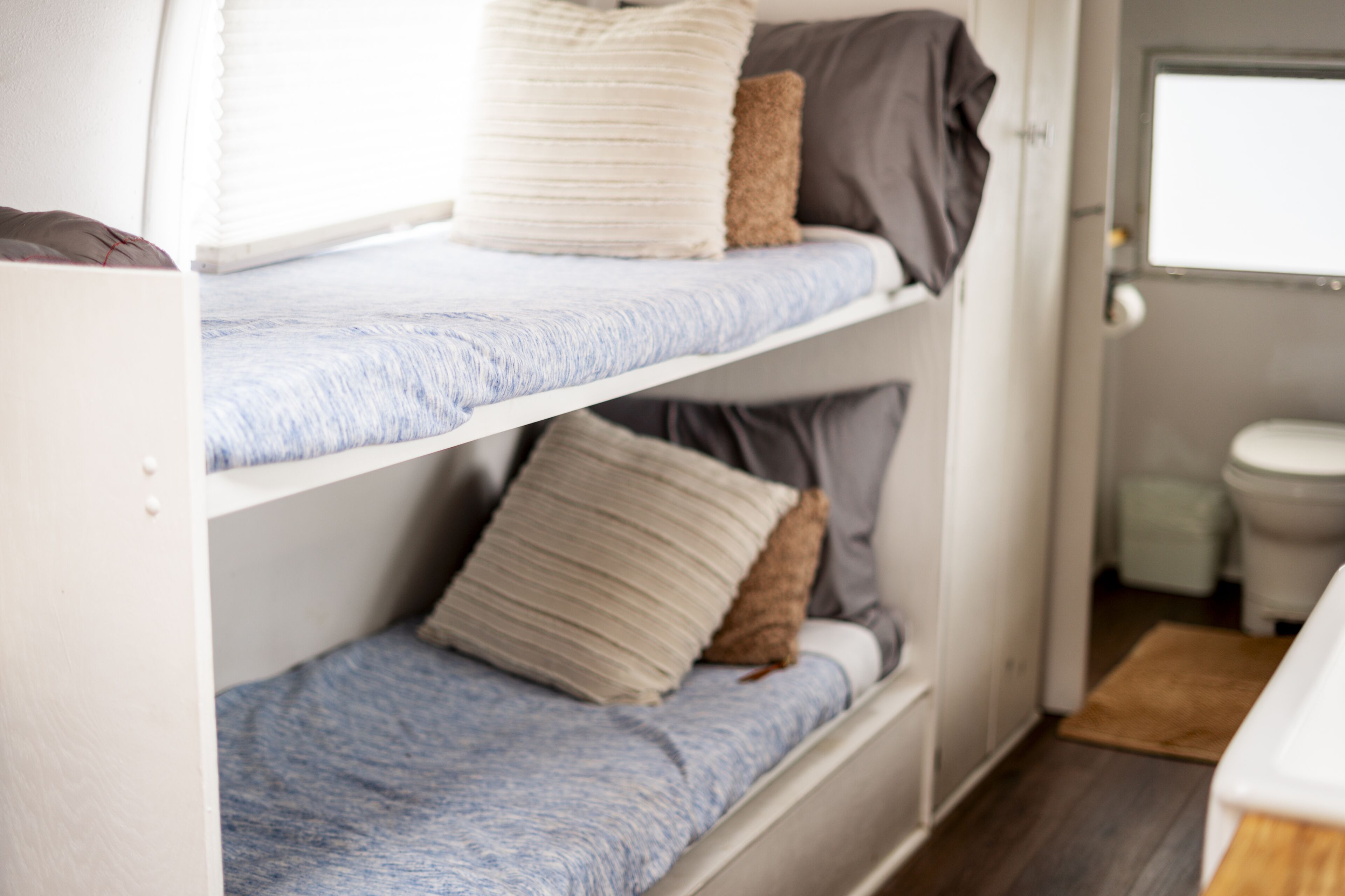 A bunk bed with two levels, each with a pillow and a blanket. There is a window above the bed and a door leading to a bathroom.