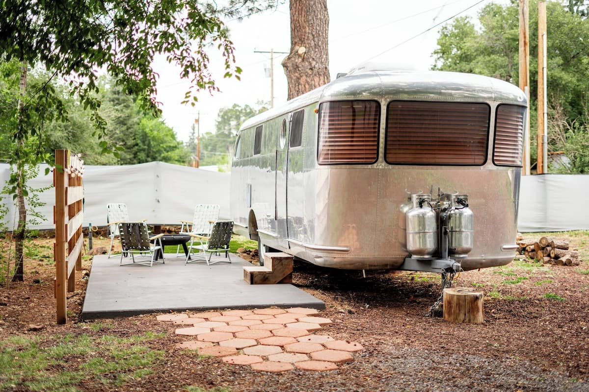 Ein Wohnwagen im Vintage-Stil mit Grill und zwei Propangasflaschen, geparkt auf einem Kiesplatz mit gepflasterter Terrasse und Außenmöbeln.