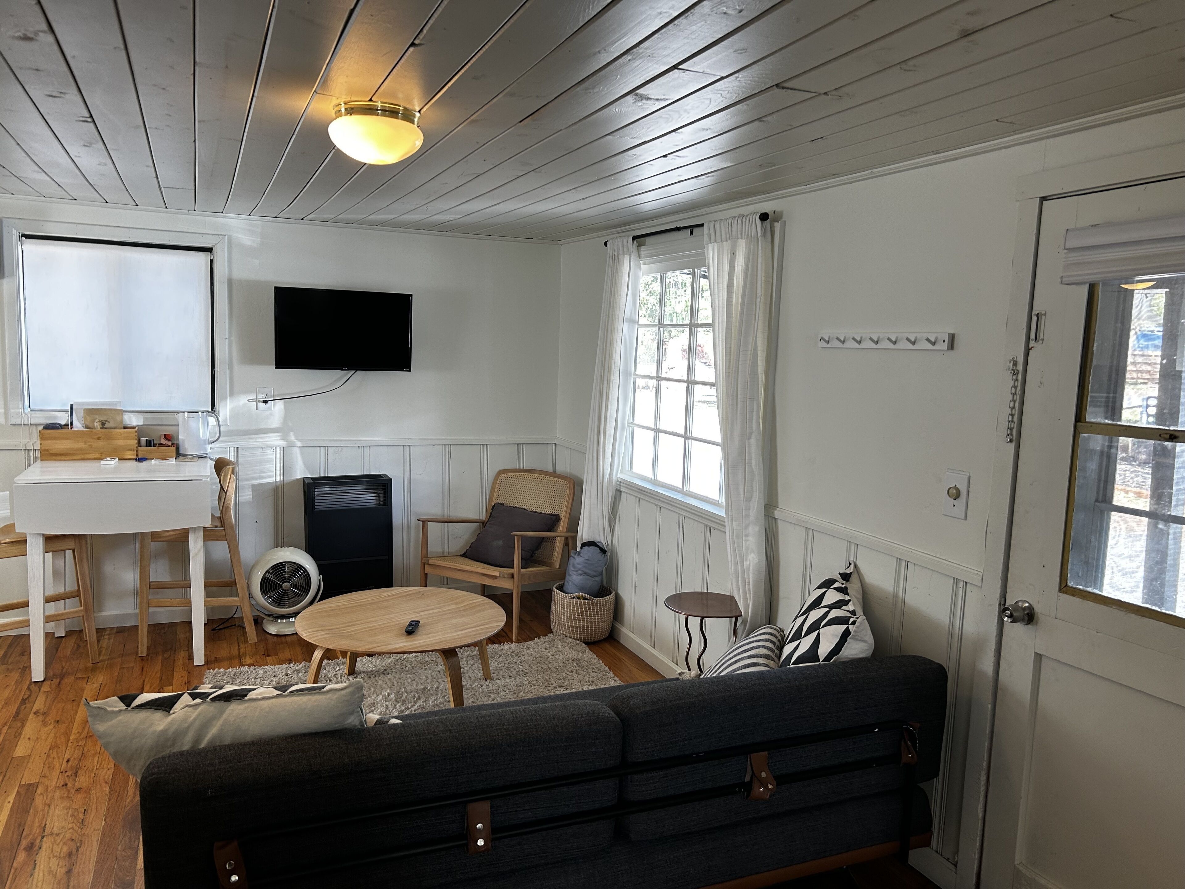 Ett vardagsrum med en soffa, ett litet runt bord, en tv som är monterad på väggen, ett fönster med gardiner och en dörr.