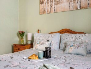 Una cama bien hecha con ropa de cama floral, una mesita de noche con un jarrón de flores, una lámpara y una bandeja con un croissant y una taza.