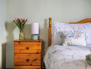 Un dormitorio con una mesita de noche de madera, un jarrón con tulipanes rosados, una lámpara blanca y una cama con ropa de cama estampada.