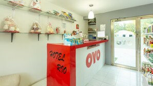 Un espace d’accueil d’hôtel avec un comptoir rouge, des étagères remplies de divers objets et une vue sur l’extérieur à travers une porte vitrée.