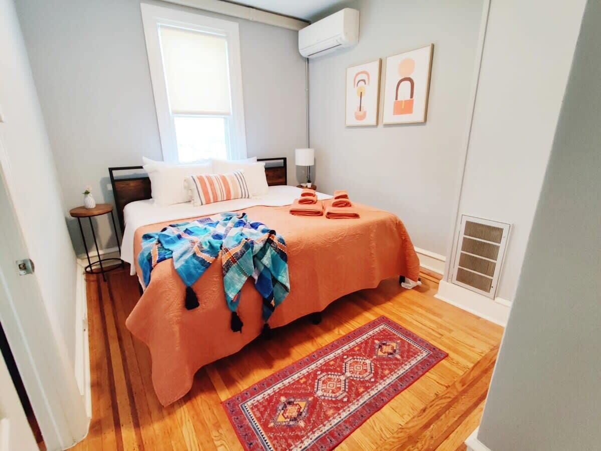 A bedroom with a bed, a nightstand, a lamp, a window, and a red rug.