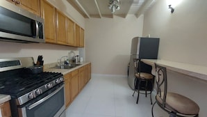 A compact kitchen with wooden cabinets, a stainless steel stove, a microwave, and a refrigerator. There are two bar stools and a small table. - The Golden Gate Inn (Basseterre)