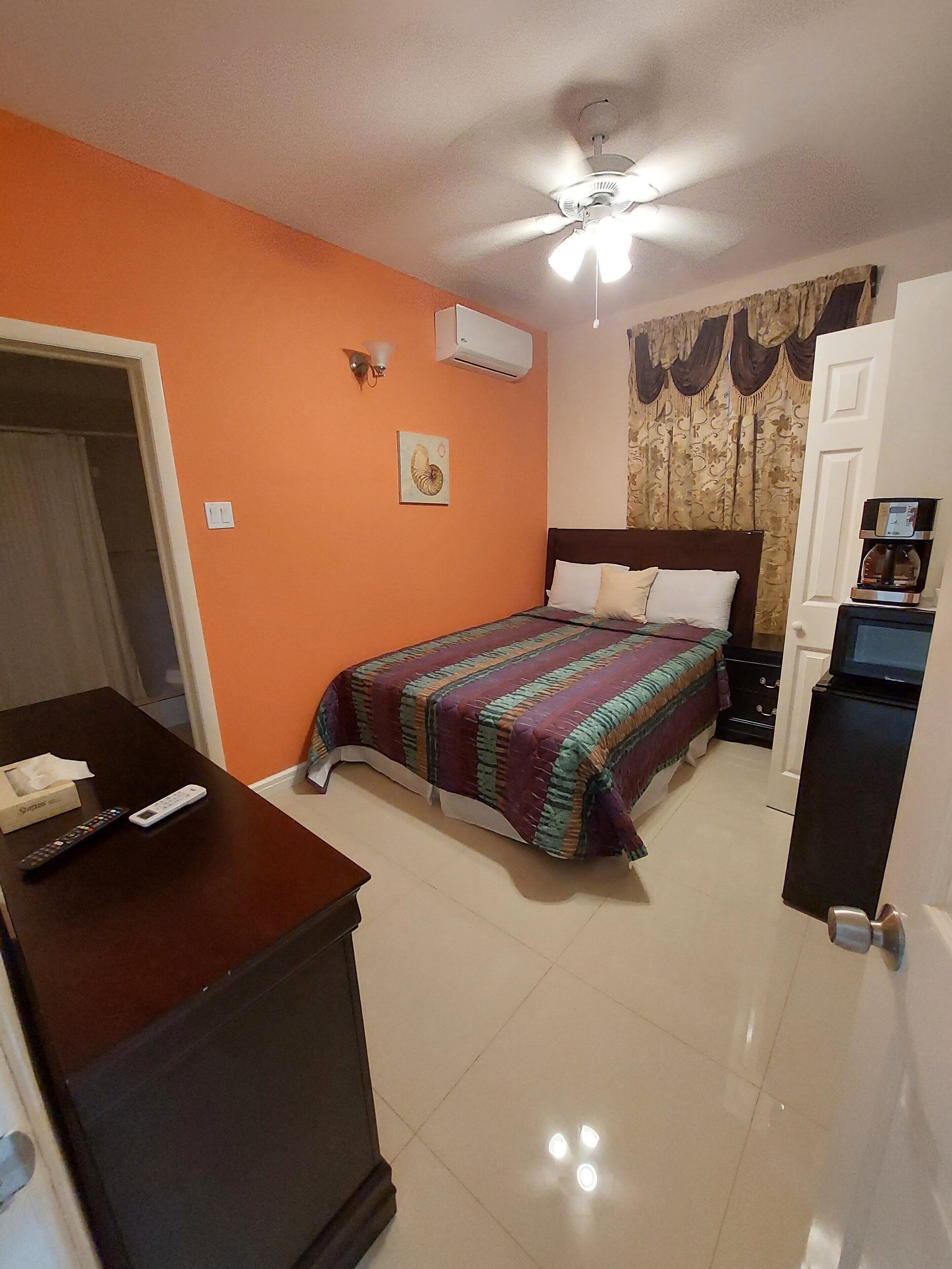 Une chambre d’hôtel avec un mur orange, un lit recouvert d’une courtepointe à motifs, un bureau avec une télécommande, une machine à café et un ventilateur de plafond.