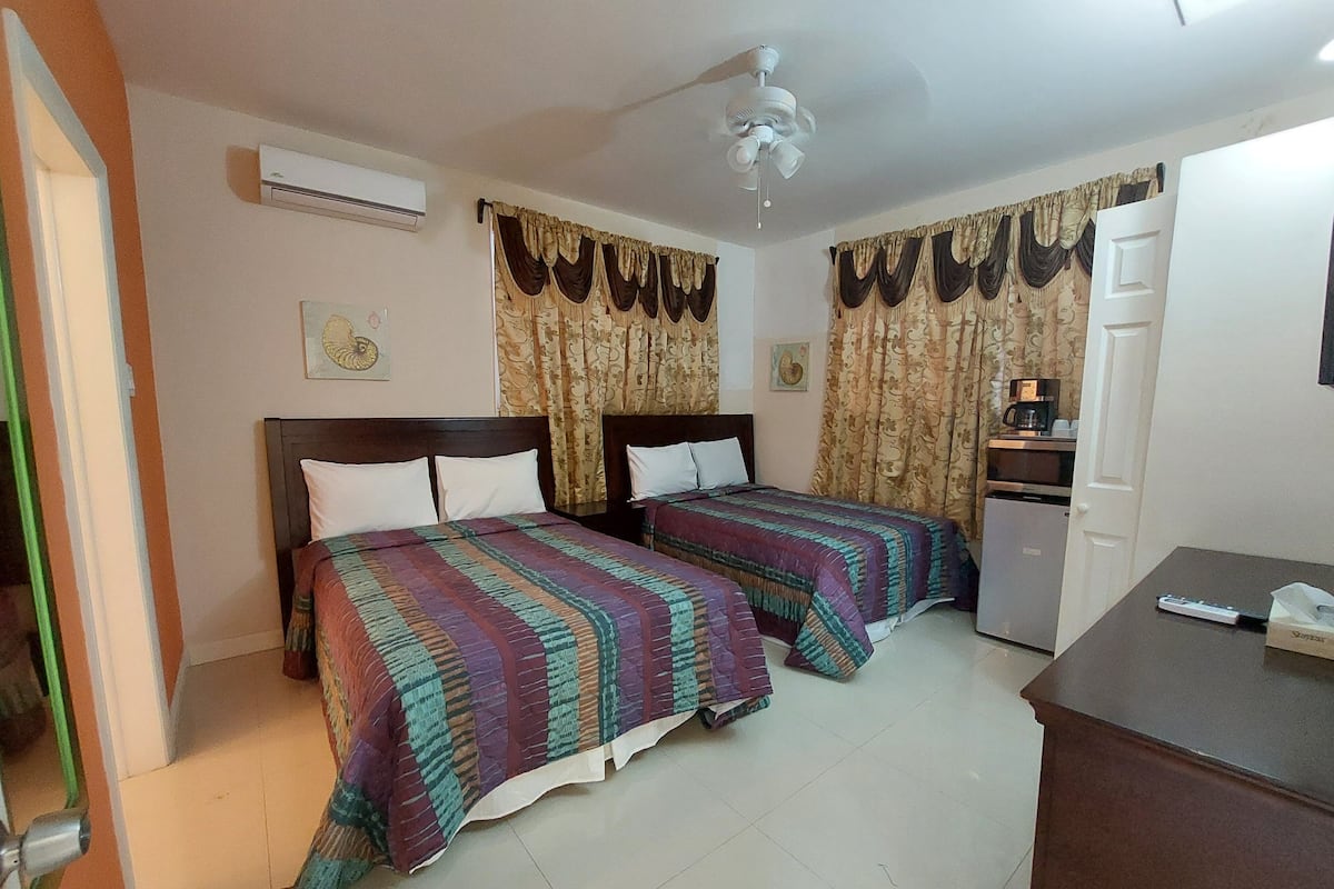 A hotel room with two beds, a ceiling fan, an air conditioner, curtains, a coffee machine, and a desk with a remote control.