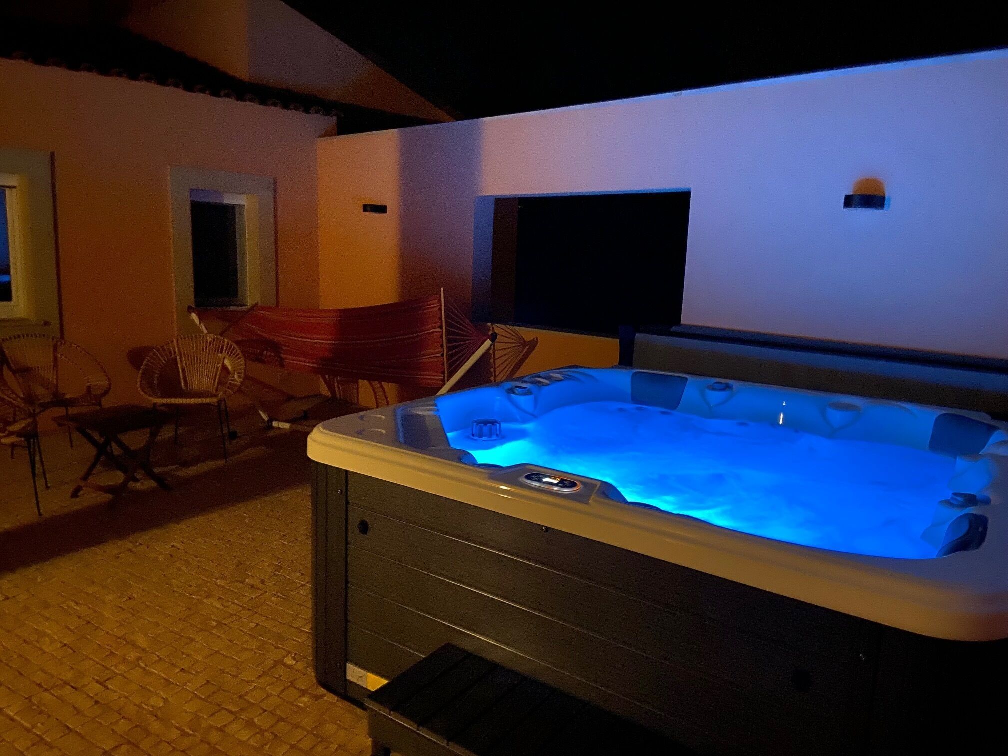 A hot tub with a control panel, surrounded by outdoor chairs and a TV screen mounted on the wall.