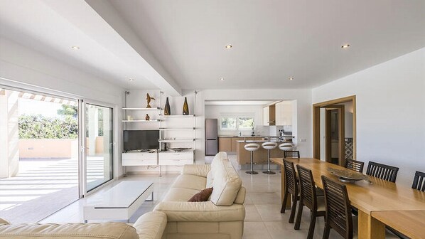 A modern living room with a sofa, a dining table with chairs, a TV, and a kitchen in the background.