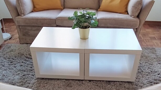 Un salón con un sofá beige, dos cojines amarillos, una mesa de centro blanca y una planta en maceta.
