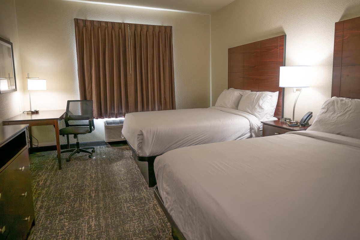 A hotel room with two beds, a desk, a chair, a lamp, and an air conditioning unit.