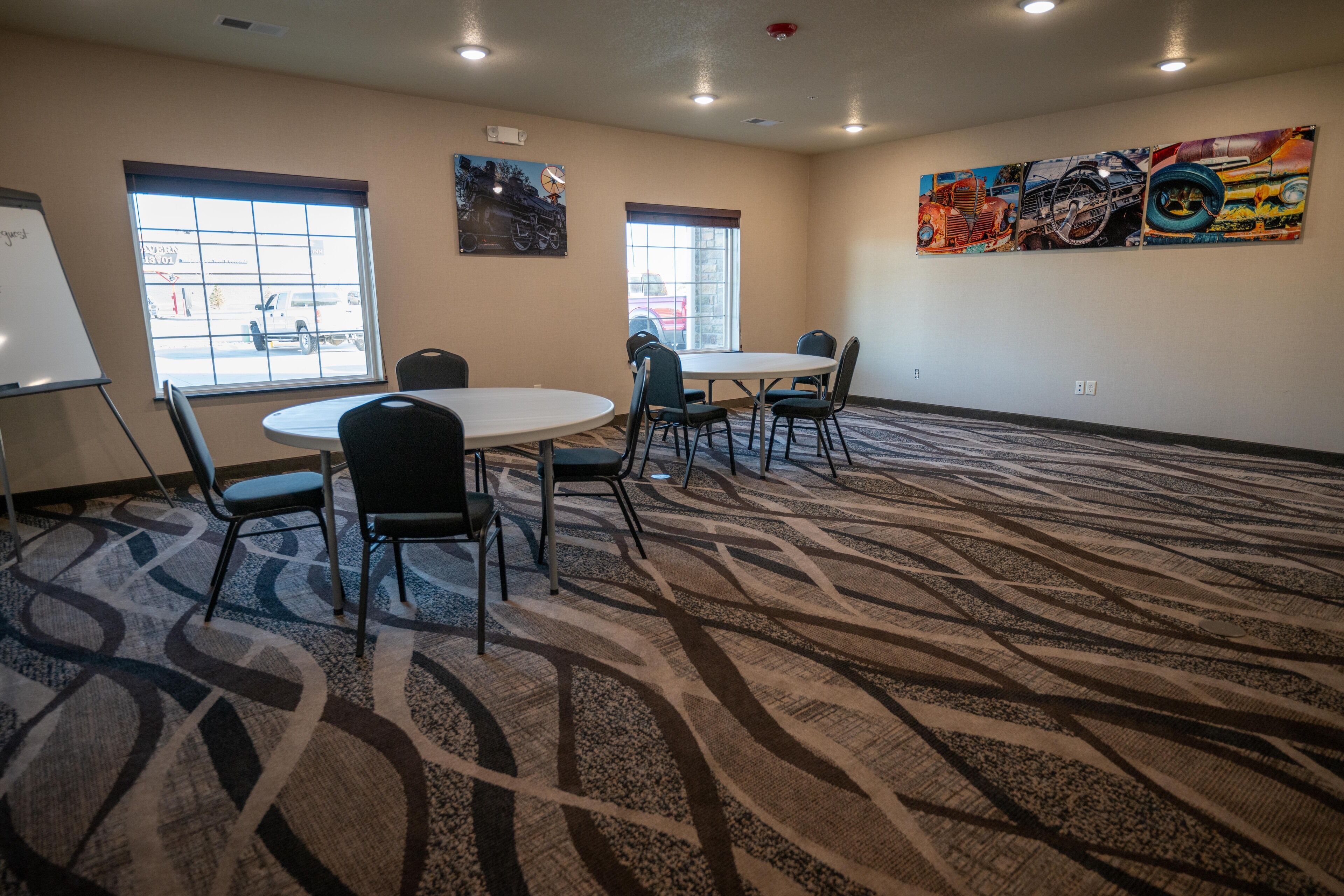 A conference room with a round table, chairs, a whiteboard, and windows with a view of parked cars.