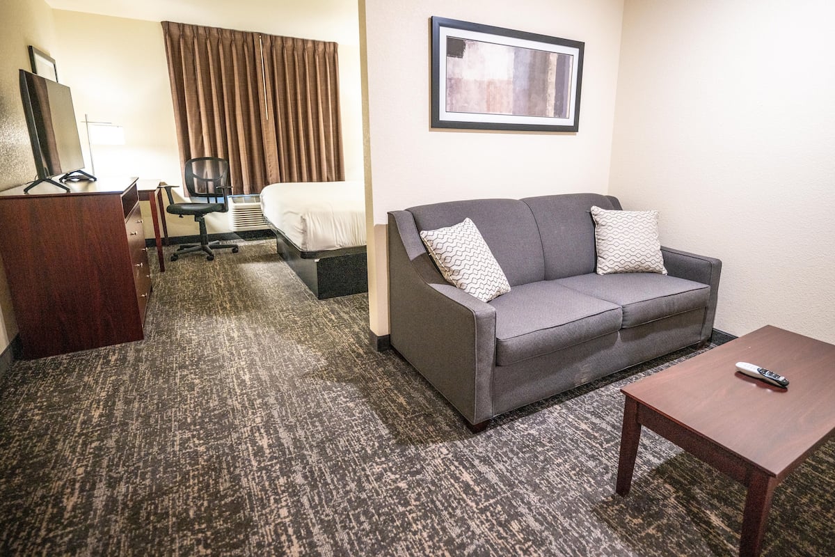A hotel room with a grey sofa, a wooden coffee table, a desk with a chair, a bed, and a framed picture on the wall.