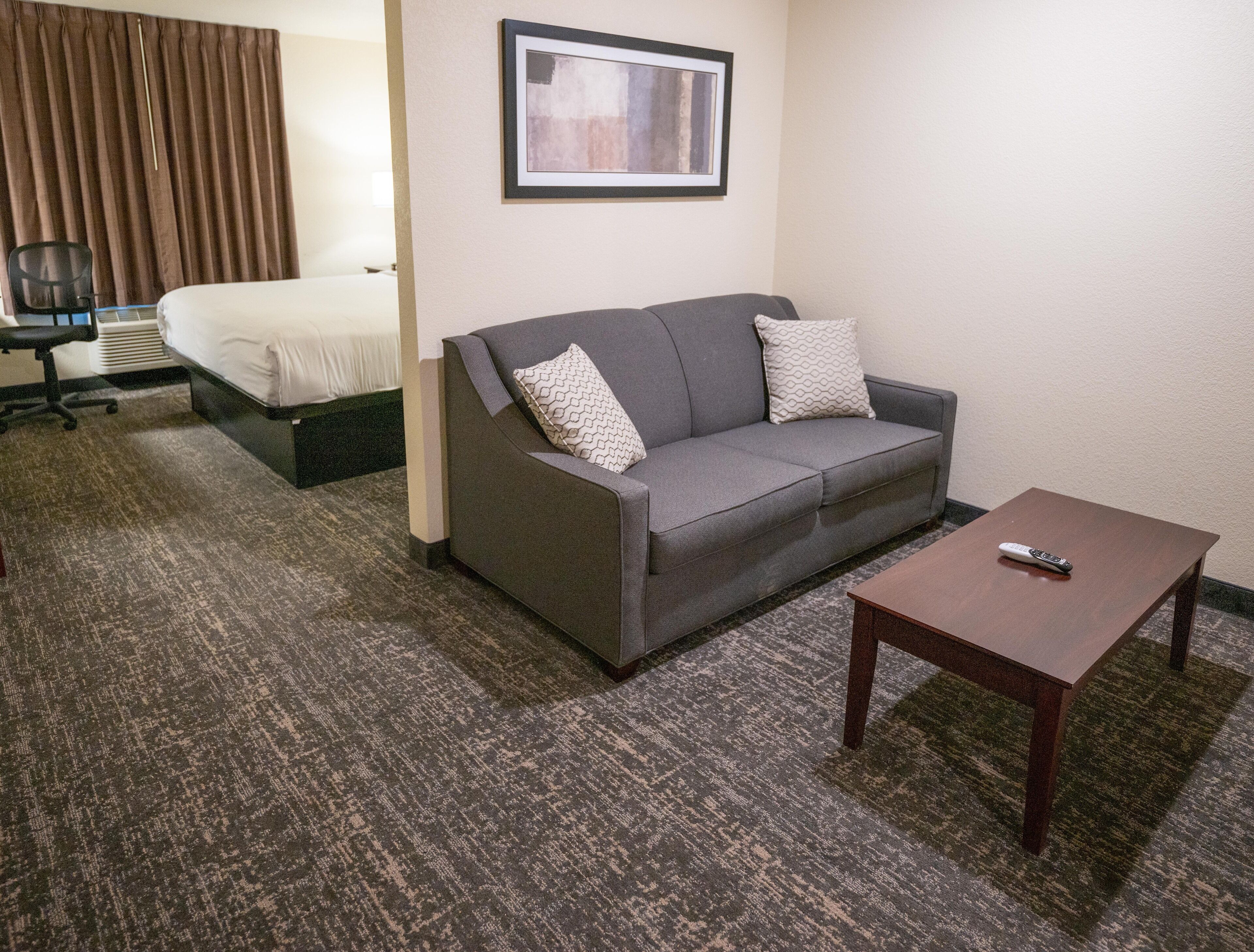 A hotel room with a gray sofa, a wooden coffee table, a bed with white bedding, a framed picture on the wall, and a chair.