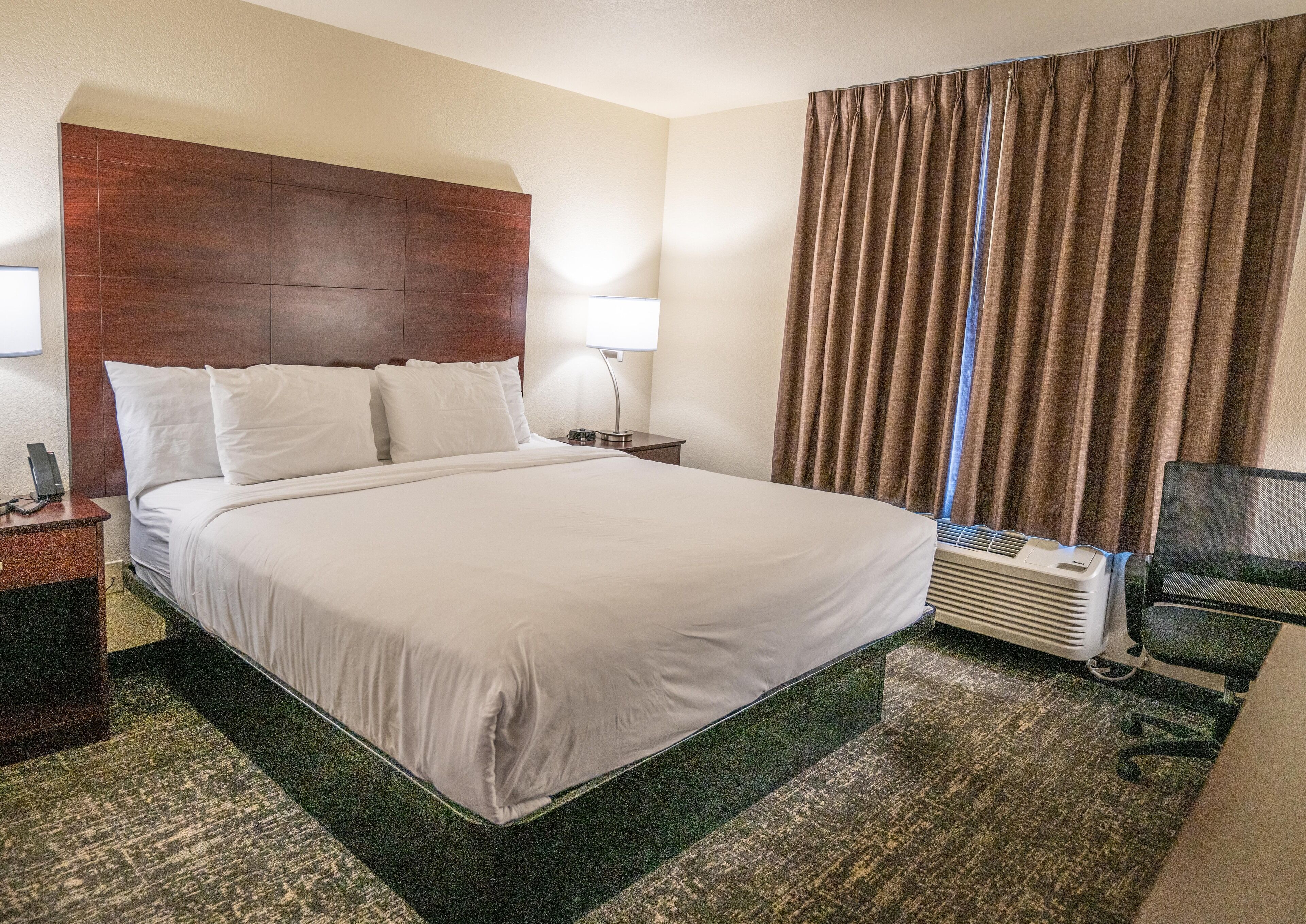 A hotel room with a large bed, a wooden headboard, a desk with a lamp, a chair, and a window with curtains.