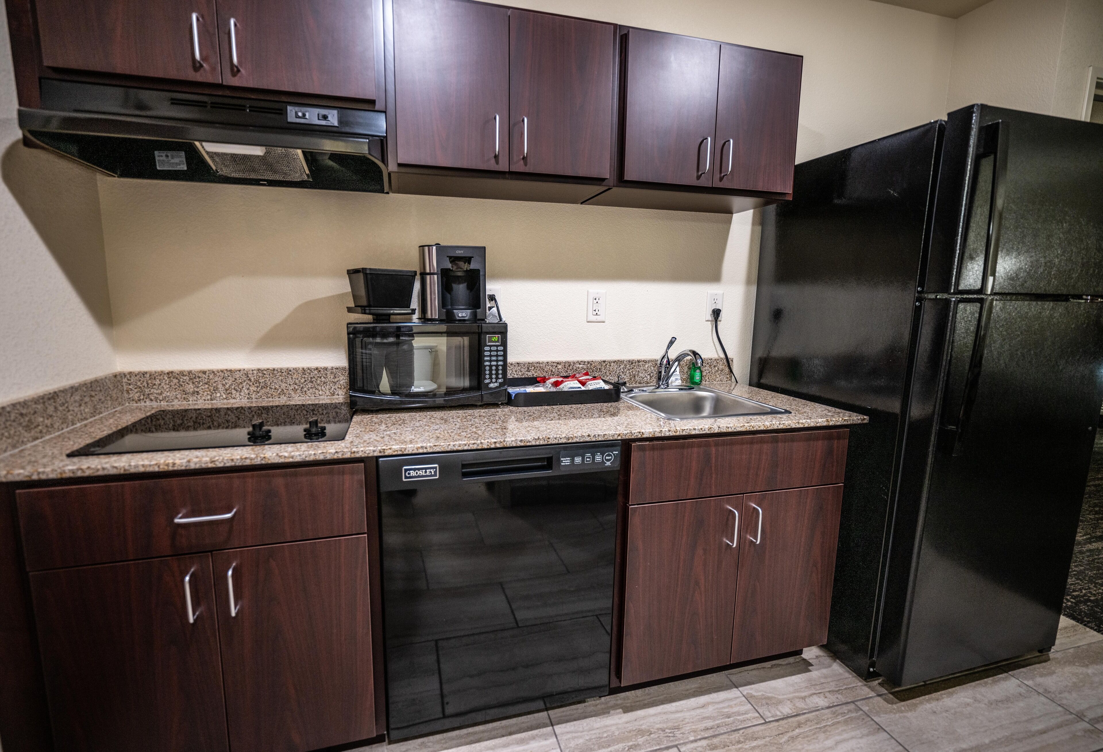 A kitchen with dark wood cabinets, a black refrigerator, a built-in dishwasher, a microwave, and a coffee maker.
