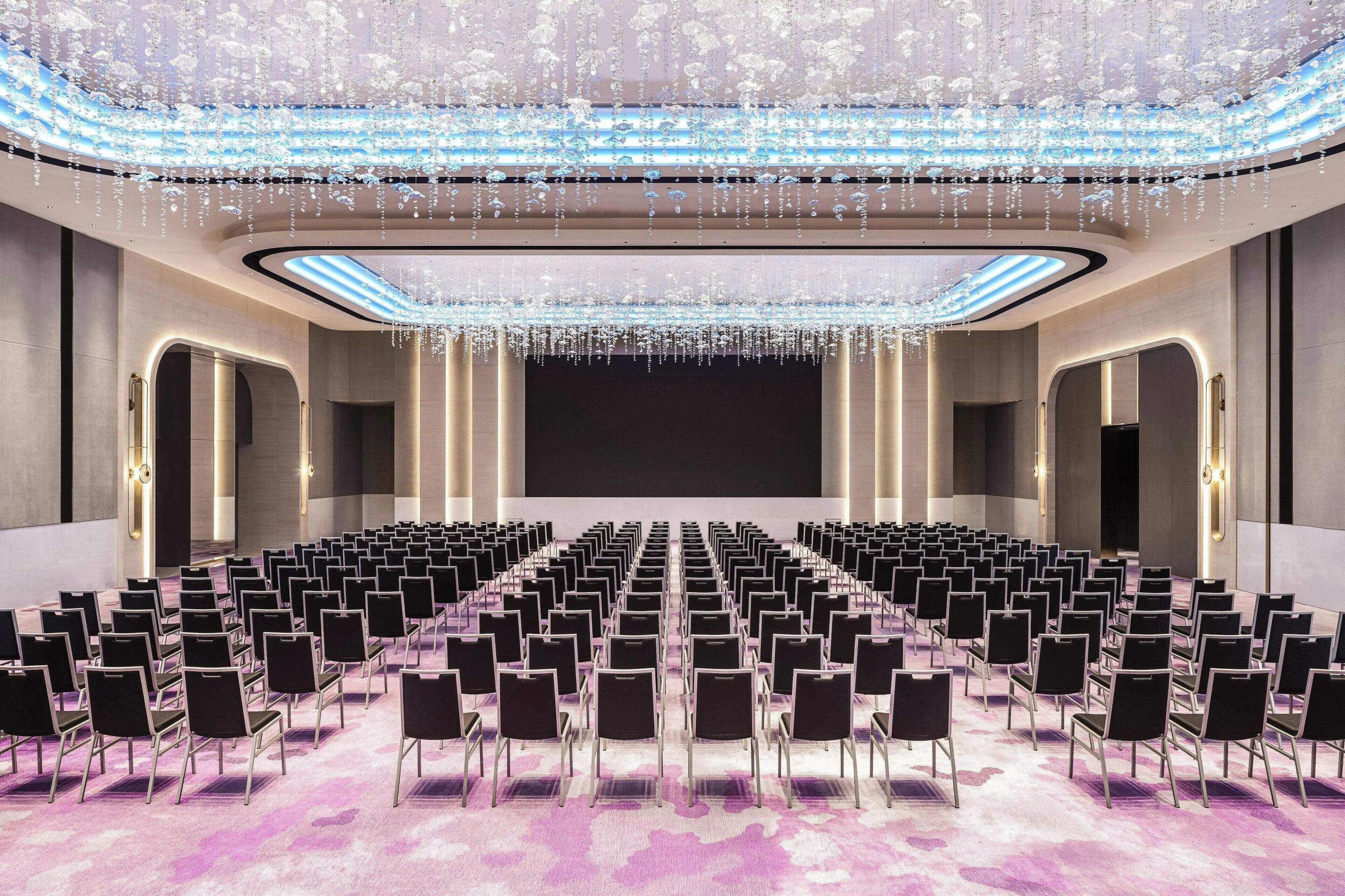 Uma sala de conferências ampla, com fileiras de cadeiras, palco e teto com luminárias pendentes.