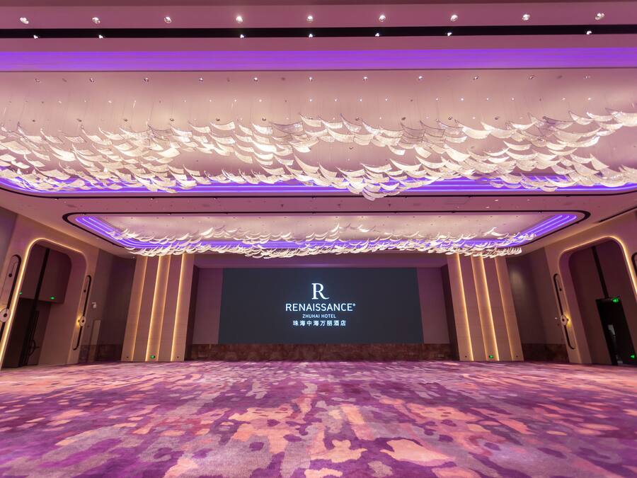 A spacious conference room with a large screen displaying the hotel name, a patterned carpet, and a ceiling with a unique light fixture.