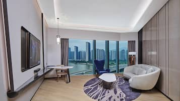 Habitación de hotel moderna con un ventanal amplio que ofrece vistas a la ciudad, un televisor de pantalla plana, una mesa de centro redonda y una zona de estar acogedora.
