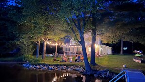 Un espace extérieur aménagé avec des sièges, situé au bord d’un lac, avec une maison en arrière-plan, entourée d’arbres et illuminée par une guirlande lumineuse.